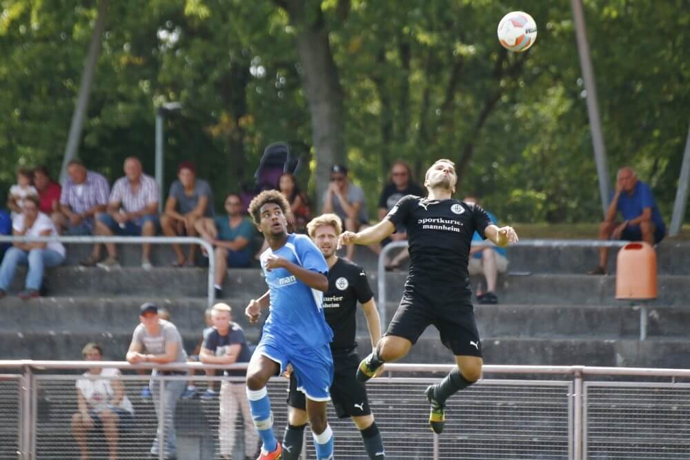 Heddesheims Anthony Loviso kommt vor seinem Dossenheimer Gegenspieler an den Ball und köpft ihn weg. Im Hintergrund Daniel Gulde (schwarzes Trikot). Bild: Rafael Kowollik.